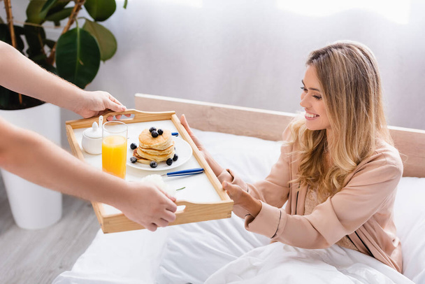 Mann reicht Frühstückstablett mit Pfannkuchen und Orangensaft neben lächelnder Freundin im Bett  - Foto, Bild
