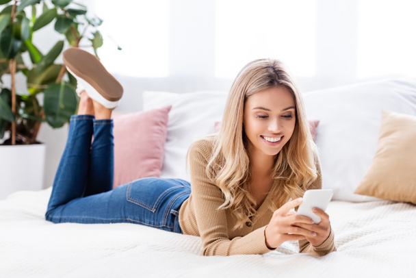 Cheerful woman using smartphone while lying on bed at home  - Photo, Image