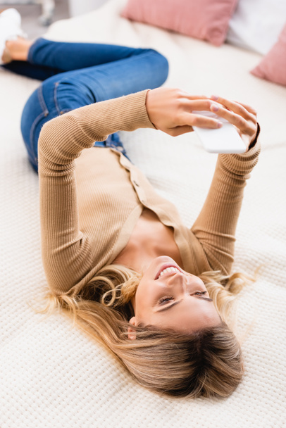 Улыбающаяся женщина с помощью смартфона на кровати дома  - Фото, изображение