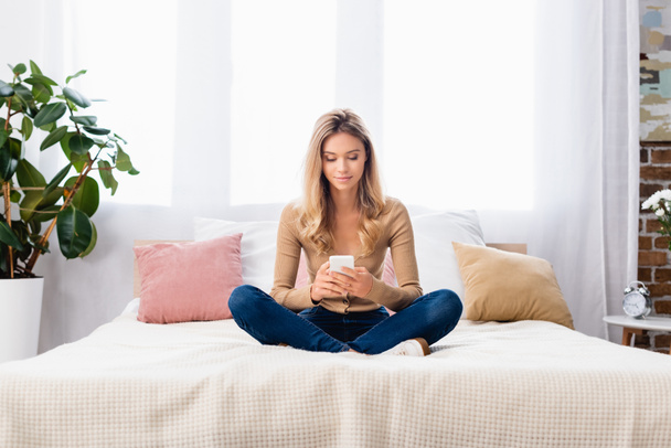 Young woman with crossed legs using smartphone on bed at home  - Φωτογραφία, εικόνα