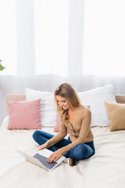 Кузнечиха работает на ноутбуке, сидя дома на кровати  - Фото, изображение