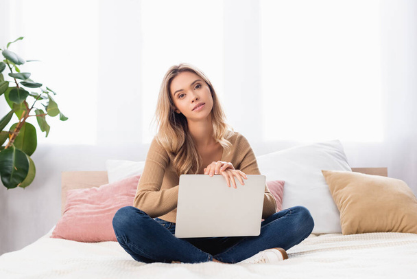 Young woman looking at camera while holding laptop in bedroom  - Photo, image
