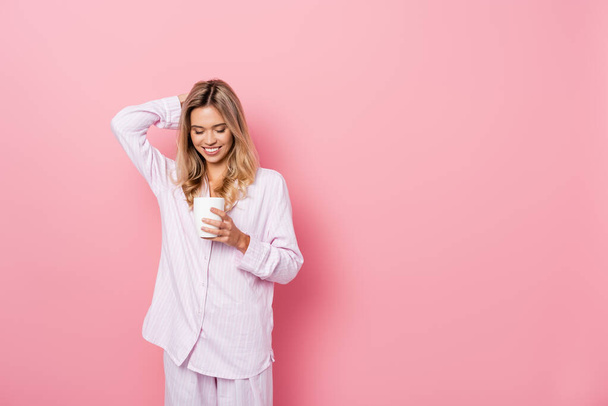 Χαμογελαστή γυναίκα με πιτζάμες με το χέρι κοντά στο κεφάλι κρατώντας κύπελλο σε ροζ φόντο - Φωτογραφία, εικόνα