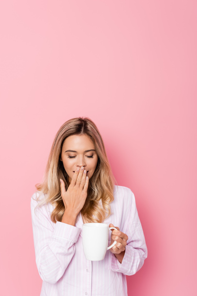 Young woman in pajamas yawning and holding cup on pink background - Photo, image