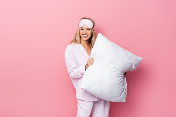 Young woman in pajamas and blindfold holding white pillow on pink background - Photo, image