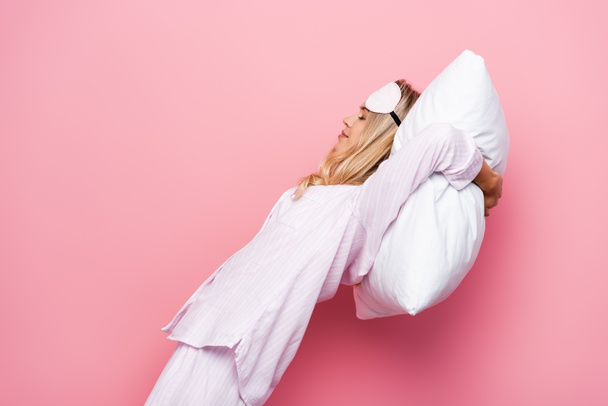 Young woman in pajamas and blindfold holding pillow near head on pink background - Foto, Imagem