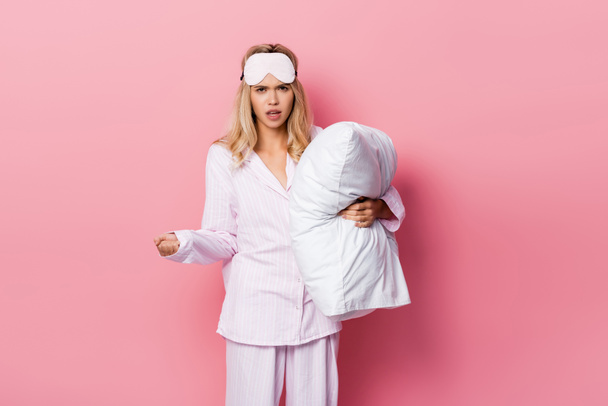 Mujer enojada en pijama y con los ojos vendados sosteniendo almohada sobre fondo rosa - Foto, Imagen