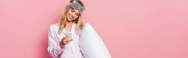 Smiling woman in blindfold and pajamas holding pillow and using smartphone on pink background, banner  - Fotoğraf, Görsel