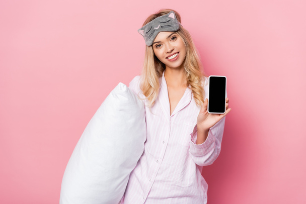Cheerful woman in blindfold and pajamas holding pillow and showing smartphone with blank screen on pink background - Φωτογραφία, εικόνα