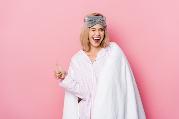 Mujer alegre en pijama y manta guiñando y mostrando como sobre fondo rosa - Foto, Imagen