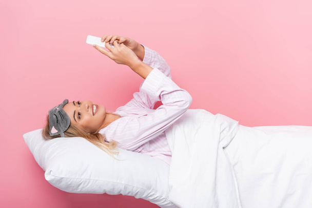 Smiling woman in blindfold using smartphone on bedding on pink background - 写真・画像