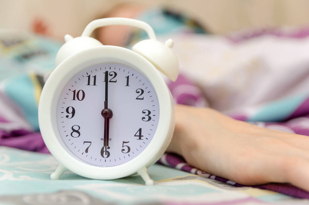 reloj despertador blanco está en la cama junto a la mano de una mujer, madrugando - Foto, imagen