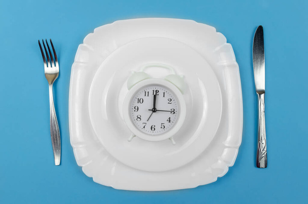 relógio de alarme branco está em uma placa redonda branca, que está em uma placa branca maior, com uma faca à direita e um garfo à esquerda em um fundo azul - Foto, Imagem