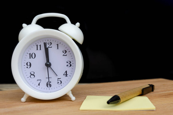 white alarm clock stands on a wooden table with a writing sheet and a pen against a black wall, copy space - Photo, Image