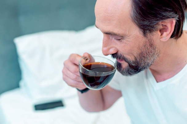 Positive Caucasian man smiling and taking a sip of coffee from a glass cup - Fotó, kép