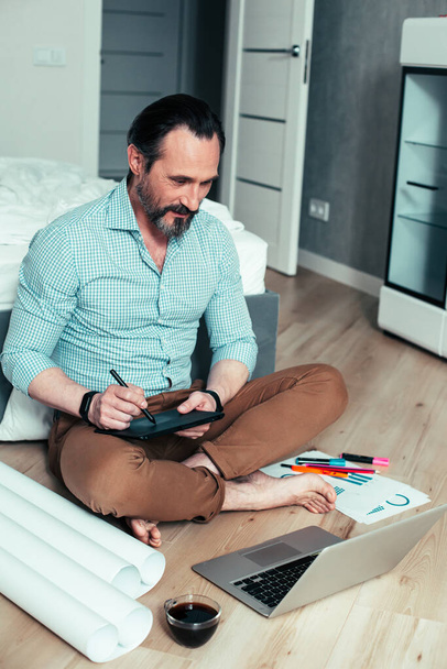 Веселий молодий чоловік сидить на підлозі в спальні і дивиться на екран ноутбука під час малювання
 - Фото, зображення