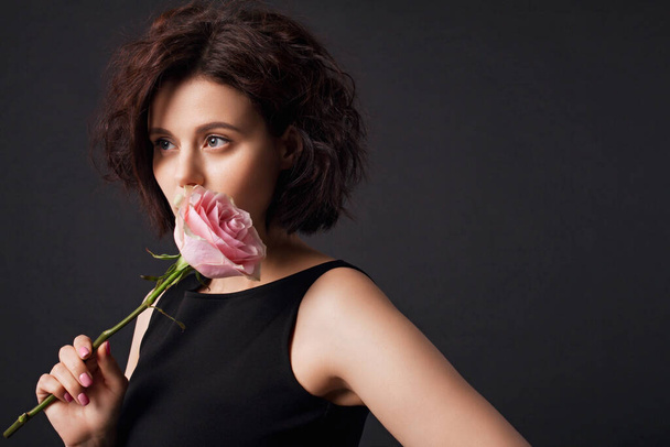 Young beautiful woman holding a pink rose. Brunette girl with short curly hair in a black dress. Black background. - Zdjęcie, obraz