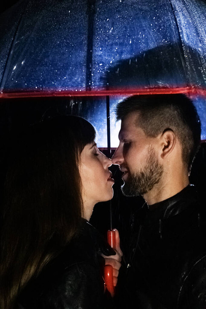 Um casal apaixonado sob um guarda-chuva beijando, e chuva está caindo sobre eles. Fotografia noturna de pessoas. O conceito de proteger relacionamentos e amor longo para um casal. - Foto, Imagem