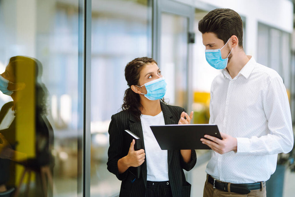 Two colleagues in protective face masks in modern office discussing together work issues. Business People back at work in office after quarantine. Covid-19. - Photo, Image