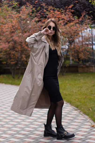 привлекательная стройная юная брюнетка в модном окопе, черном платье и солнечных очках в парке. осень, желтые листья - Фото, изображение