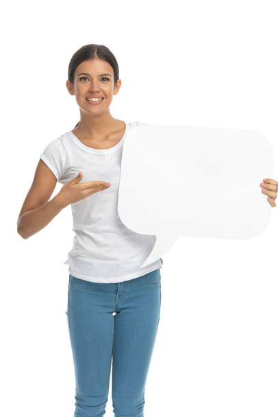 Mujer casual positiva sonriendo y presentando burbuja del habla mientras está de pie en el fondo del estudio blanco - Foto, imagen