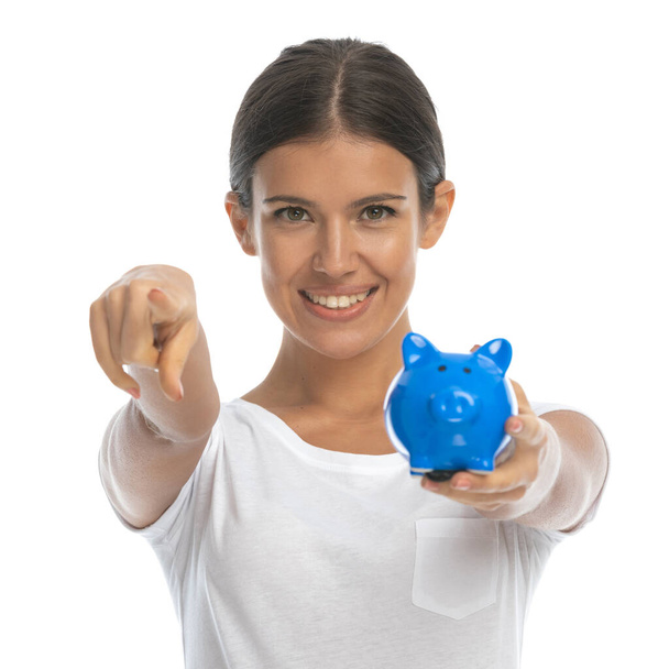glimlachende casual vrouw holding piggy bank en wijzen terwijl staan op witte studio achtergrond - Foto, afbeelding