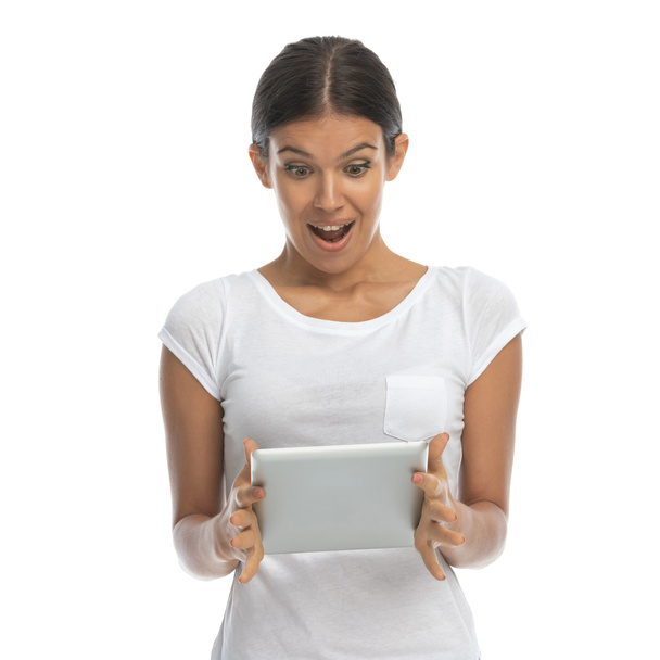 Возбужденная случайная женщина смеется и задыхается, держа планшет, стоя на белом фоне студии - Фото, изображение