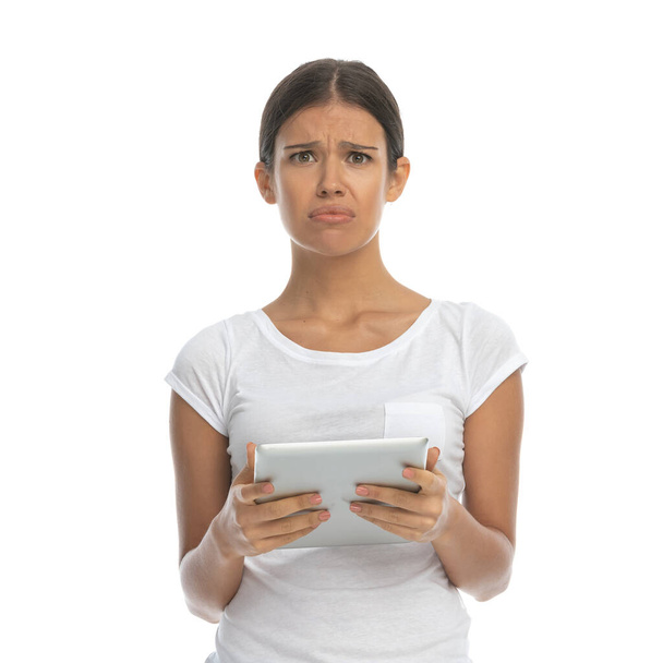 Molesto mujer casual sosteniendo la tableta mientras está de pie sobre fondo de estudio blanco - Foto, Imagen