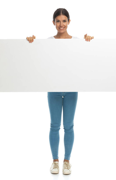 Happy casual woman covering her body with blank billboard while standing on white studio background - Foto, imagen