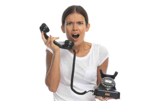 Έκπληκτη περιστασιακή γυναίκα μιλάει στο παλιό τηλέφωνο και λαχανιάζει ενώ στέκεται σε λευκό φόντο στούντιο - Φωτογραφία, εικόνα