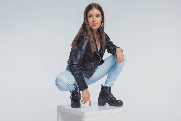Confident fashion model smiling, wearing leather jacket while crouching on gray studio background - Photo, Image