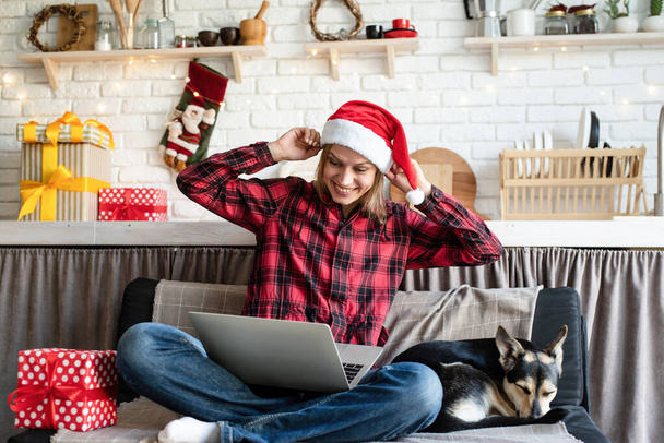Chhristmas pozdrowienia online. Szczęśliwa młoda kobieta w kapeluszu Mikołaja pozdrawiając przyjaciół w rozmowie wideo na laptopie pokazując prezent świąteczny - Zdjęcie, obraz