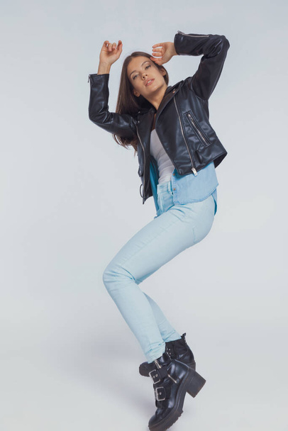 Charming fashion model adjusting her hair while wearing leather jacket and dancing on gray studio background - Fotoğraf, Görsel