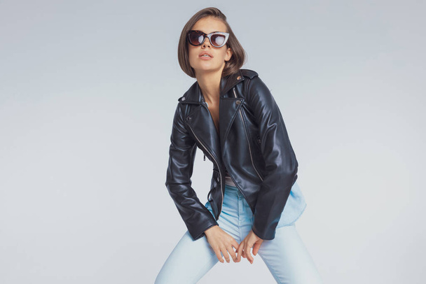 Sebevědomý módní model pózující v kožené bundě a slunečních brýlích, stojící na šedém pozadí studia - Fotografie, Obrázek
