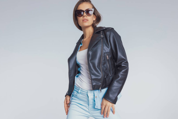 Modèle de mode confiant portant une veste en cuir et des lunettes de soleil, debout sur fond de studio gris - Photo, image