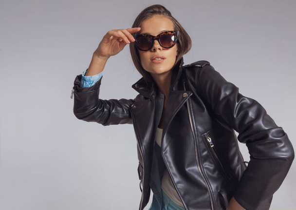 Selbstbewusstes Mode-Model blickt in die Ferne, während es in Lederjacke und Sonnenbrille vor grauem Studio-Hintergrund steht - Foto, Bild
