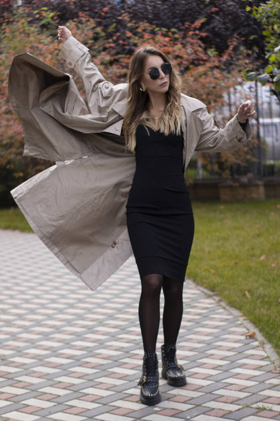 ファッショナブルなトレンチで魅力的なスリム若いブルネット、公園で黒のドレスとサングラス。秋黄色の葉 - 写真・画像