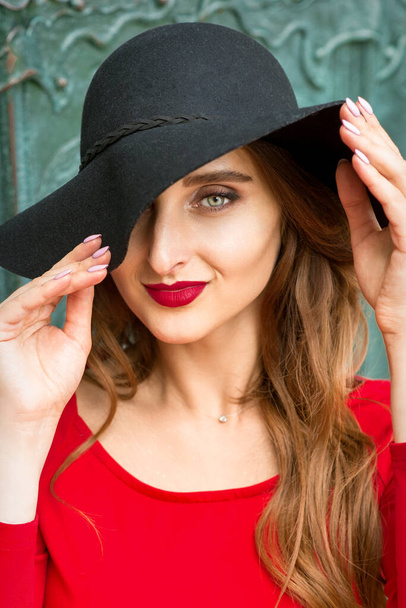 Portrait of beautiful fashionable caucasian young woman in red dress wearing black hat looking at camera - Foto, Imagen