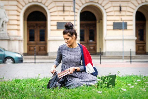 Νεαρή ενήλικη φοιτήτρια βάζει τα βιβλία σε σακίδιο κάθεται στο γρασίδι κοντά στο κτίριο του πανεπιστημίου - Φωτογραφία, εικόνα