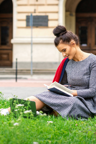 Junge kaukasische Studentin liest auf der Straße im Gras neben der Universität das Buch - Foto, Bild