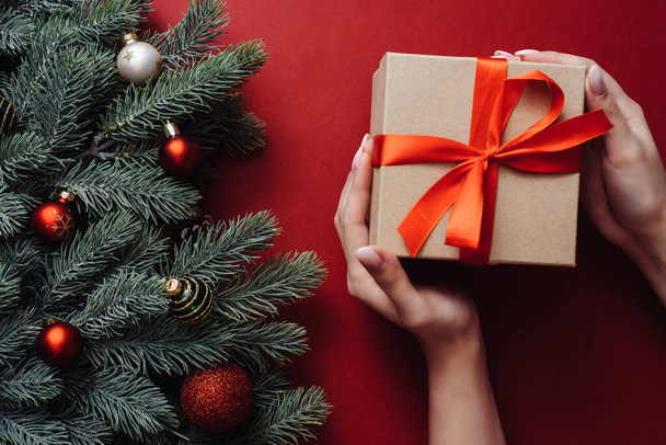Bayram hediyesi olarak Firavun dalları ve toplarıyla kırmızı arka planda Noel hediyesini tutan kadın eller. Xmas ve Mutlu Yıllar kartı. Düz yatış, üst görünüm - Fotoğraf, Görsel