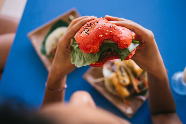 Female customer eating tasty hearty American burger with tomato bread rolls and vegan meat, hungry woman enjoying leisure time for have lunch with delicious caloric hamburger during cheat meal on diet - Photo, Image
