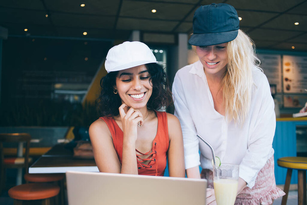 Happy women with black and blond hair in caps and casual clothes using laptop to prepare for exam in cozy cafe workspace - Photo, Image