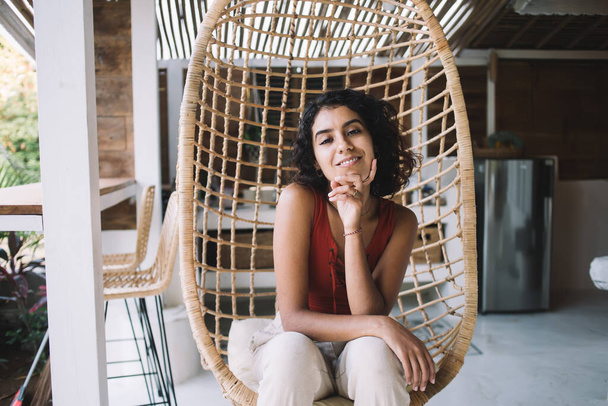 Young woman with curly hair sitting propping head in wicker chair suspended from ceiling on weekend in cozy apartment and looking at camera - Foto, Imagen