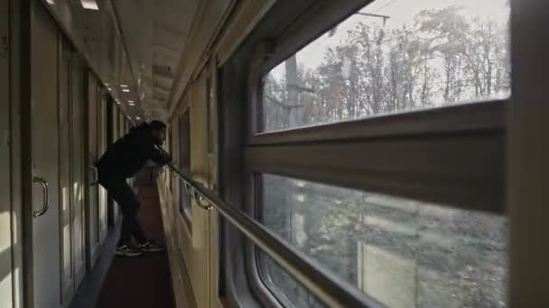 Un hombre joven y guapo se para en la ventana del tren. La vista fuera de la ventana del tren. Mira a lo lejos. Un hombre de pelo largo y barba, un turista de viaje. - Metraje, vídeo