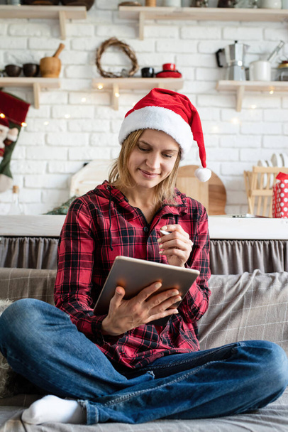Vánoční online pozdravy. mladá blondýnka v santa klobouk pracuje na tabletu sedí na gauči - Fotografie, Obrázek