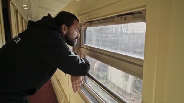 Un hombre joven y guapo se para en la ventana del tren. La vista fuera de la ventana del tren. Mira a lo lejos. Un hombre de pelo largo y barba, un turista de viaje. - Metraje, vídeo