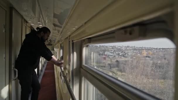 Młody, przystojny mężczyzna stoi przy oknie pociągu. Widok za oknem pociągu. Spójrz w oddali. Mężczyzna z długimi włosami i brodą, turysta na wycieczce. - Materiał filmowy, wideo