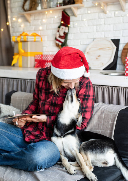 Christmas online greetings. young blond woman in santa hat working on tablet sitting on the couch with her dog - Photo, Image