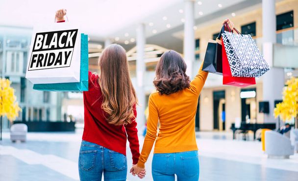 Просмотр фотографии двух молодых женщин в разноцветной одежде с сумками в руках с отпечатками черной пятницы. - Фото, изображение